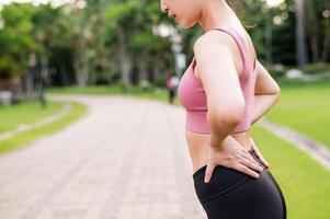 Woman jogger. young 30s asian female wearing pink sportswear holding her back and hip pain after running exercise in public park. Pain in activity concept. photo