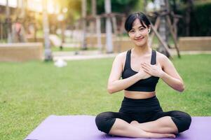 Portrait fit cheerful young asian woman 30s wearing black sportswear holding hands on chest while listening relax music and sitting on yoga mat in public park. Healthcare wellness concept. photo