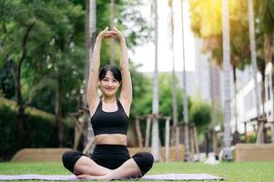 Portrait fit young asian woman 30s wearing black sportswear listen relaxing music while stretching muscle before yoga exercise on yoga mat in green nature park. wellbeing concept. photo