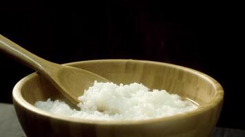 Close up for steamed rice in a wooden bowl with wooden stirring spoon, isolated on black background. Freshly cooked rice with steam in deep, brown plate with big spoon, food, cooking concept. video