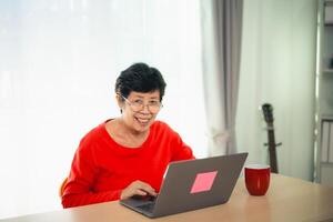 mayor antiguo asiático mujer trabajando después Jubilación utilizando ordenador portátil a hogar. antiguo persona de libre dedicación trabajando o aprendizaje nuevo tecnología en ordenador portátil en vivo habitación. Jubilación actividad concepto. foto