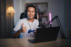 Asian influencer man wearing glasses smile cheerful Thumb up using joystick playing game live streaming greeting video conference with laptop and wearing headphone. Asian man teaching online concept. photo