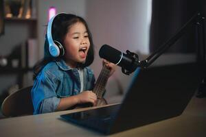 asiático joven niña sonriente En Vivo transmisión actuación jugando ukelele y canta un canción. asiático niña aprendizaje guitarra y canto en línea. músico grabación música con ordenador portátil y jugando acústico guitarra. foto