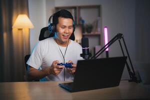 Asian influencer man wearing glasses smile cheerful using joystick playing game live streaming greeting video conference with laptop and wearing headphone. Asian man teaching online concept. photo