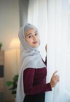 retrato de hermosa asiático musulmán mujer en un hasta los hombros hijab en pie lado ventana en el vivo habitación con grande ventanas oriental mujer sonriente brillantemente posando en un moderno hogar. foto