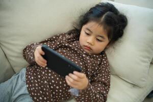 Asian child girl serious or concentrate and using mobile phone screen on couch sofa. Baby smiling funny time use mobile phone. Too much screen time. Girl watching videos while tv, Internet addiction. photo