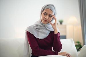 Muslim islam Asian woman wearing hijab depressed sadly serious  having stressful depression sad time sitting on sofa in the living room at home. Depression woman sad serios health mental concept. photo