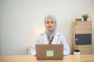 musulmán islam asiático médico hombre vistiendo hijab y estetoscopio trabajando con ordenador portátil computadora. hembra médico trabajo a hogar oficina hospital. salud hospital concepto. foto