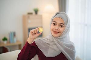 Muslim islam Asian woman wearing hijab smiling holding gold bitcoin crypto currency blockchain sitting on sofa in living room at home. Crypto currency blockchain Saving investment wealth concept. photo