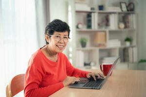 Senior old asian woman working after retirement using laptop at home. Old freelancer working or learning new technology on laptop in living room. Retirement activity concept. photo