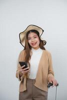 Asian smiling tourist woman with braces wearing hat and holding luggage holding mobile phone, copy space going to travel on holidays isolated on blue studio background. Travel journey concept. photo