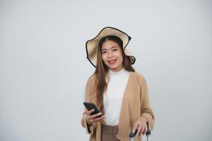 Asian smiling tourist woman with braces wearing hat and holding luggage holding mobile phone, copy space going to travel on holidays isolated on blue studio background. Travel journey concept. photo