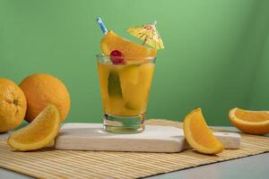 Yellow cocktail in glass glass filled with ice and mint leaves garnished with orange slice, cherry, sorbet and yellow umbrella on a bamboo napkin with orange pieces photo