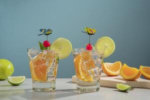 Two cocktails in glass glass filled with ice and orange slice garnished with lemon slice, cherry and butterfly pin on a white table with pieces of orange and lemon photo