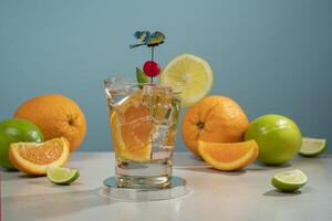 Cocktail in glass glass filled with ice and orange slice garnished with lemon slice, cherry and butterfly pin on a white table with pieces of orange and lemon photo