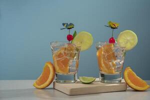 dos cócteles en vaso vaso lleno con hielo y naranja rebanada adornado con limón rebanada, Cereza y mariposa alfiler en de madera tablero con piezas de naranjas y limón en el mesa foto