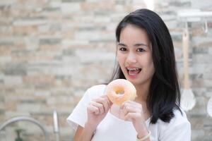 retrato de asiático mujer comiendo rosquilla en el cocina a hogar foto