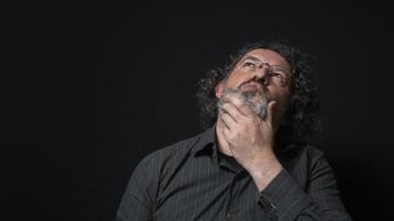 Man with white beard and black curly hair with expression of indecision and doubt, looking up, wearing black shirt against black background photo