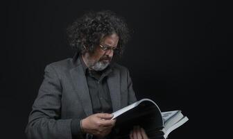 hombre con blanco barba y negro Rizado pelo leyendo un libro, vistiendo negro camisa en contra negro antecedentes foto