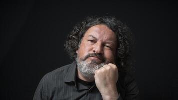 Man with white beard and black curly hair with bored expression, resting head on his hand, wearing black shirt against black background photo