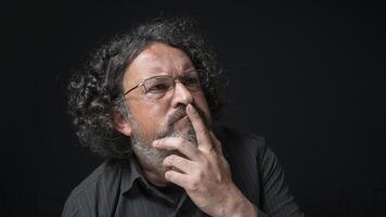 hombre con blanco barba y negro Rizado pelo con expresión de indecisión, mirando a el bien, vistiendo negro camisa en contra negro antecedentes foto