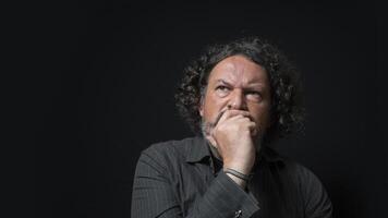 Man with white beard and black curly hair with doubtful and angry expression, with his hand on his face, wearing black shirt against black background photo