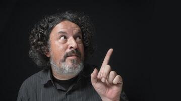 hombre con blanco barba y negro Rizado pelo con gracioso expresión, señalando arriba con su mano, vistiendo negro camisa en contra negro antecedentes foto