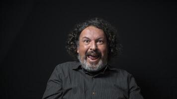 hombre con blanco barba y negro Rizado pelo con contento y emocionado expresión, vistiendo negro camisa en contra negro antecedentes foto