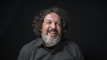 Man with white beard and black curly hair with happy expression, with half closed eyes, wearing black shirt against black background photo