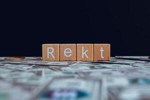 Wooden blocks with the text Rekt on a black background and crypto banknotes scattered on the ground. photo