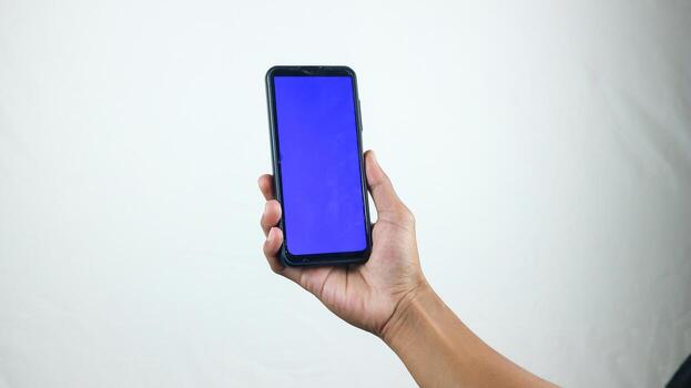 Asian man holding big smartphone with white, red, green, blue blank screen in hand, showing close to camera and pointing at device. Gadget with empty free space for mock up, banner photo