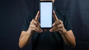 Asian man holding big smartphone with blank screen in hand, showing close to camera and pointing at device. Gadget with empty free space for mock up, banner photo