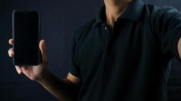 Asian man holding big smartphone with blank screen in hand, showing close to camera and pointing at device. Gadget with empty free space for mock up, banner photo