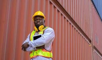Portrait of African engineer with yellow helmet and safety vest holding digital tablet while standing at cargo container yard photo