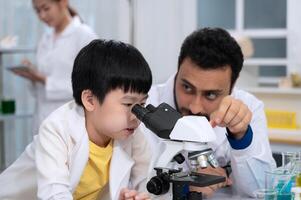 profesor con barba y estudiante vistiendo blanco laboratorio Saco en laboratorio. chico aprendizaje cómo a utilizar microscopio. foto