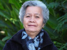 retrato de un hermosa mayor asiático mujer con blanco pelo mirando a el cámara mientras en pie en el jardín. Moda en invierno foto