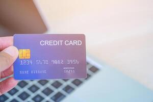 Close-up photo of the left-hand woman holding blue credit card mockup with a laptop background and copy space for text. Easy e-commerce shopping and payment by credit card