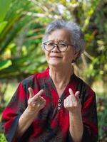 Elderly Asian woman smiling and hands making mini heart while standing in a garden photo