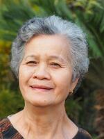 A senior Asian woman with short white hair smiling and looking at the camera while standing in a garden photo