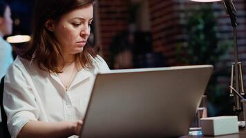 empresária pesquisando chave dados para companhia projeto, fazendo tarefas dentro escritório durante a noite. empregado olhando sobre contabilidade figuras em computador portátil tela atrasado às noite, digitando em teclado, Câmera b video