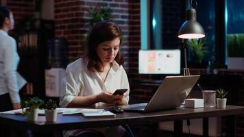 femme réponses téléphone appel de mari tandis que travail dans Bureau à ordinateur bureau. employé en utilisant téléphone intelligent à bavarder avec ami pendant Téléphone appel tandis que à travail dans de face de portable à nuit, caméra b video