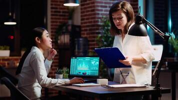 Company accountant and coworker analyzing financial data graphs on laptop screen. Employee and colleague comparing fourth quarter revenue figures in brick wall office, camera B video