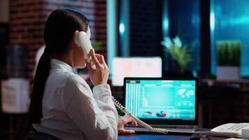 Asian employee taking landline telephone call while working with financial statistics graphs on laptop. Worker talking over the phone and solving tasks in office, doing multitasking, camera B video