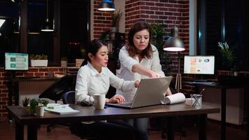 Asian businesswoman and colleague working together doing computer tasks for team project in office overnight. Coworkers looking over accounting figures on laptop screen late at night video