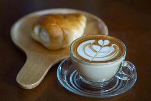 de cerca de café taza con corazón forma latté Arte espuma y un pan en de madera mesa. foto