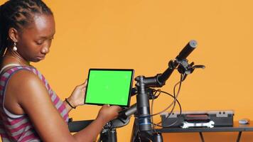 Joyful woman following tutorial videos on mockup tablet, learning to mend broken bike. Smiling cycling hobbyist having fun repairing bicycle, following online guide on green screen device, camera A