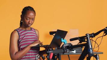 Happy worker with laptop in hand analyzing broken bike over studio background awaiting repair. Relaxed BIPOC engineer using notebook to look for replacement parts for bicycle on repair stand video