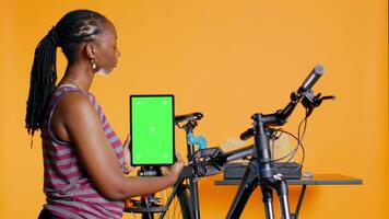 Woman holds green screen tablet, using it to advertise bike repair shop, empty placeholder for company logo, studio background. Mechanic promoting bicycle fixing service with mockup device, camera B video