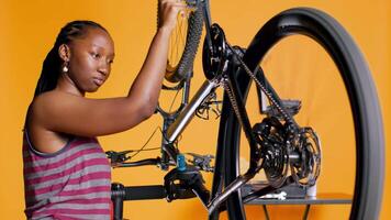 afrikanisch amerikanisch Frau Überprüfung Fahrrad Performance durch Spinnen Pedale. Fachmann gewährleisten richtig Funktion von Reifen auf Fahrrad Räder im Orange Studio Hintergrund, Kamera b video