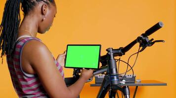 Joyful woman following tutorial videos on mockup tablet, learning to mend broken bike. Smiling cycling hobbyist having fun repairing bicycle, following online guide on green screen device, camera B